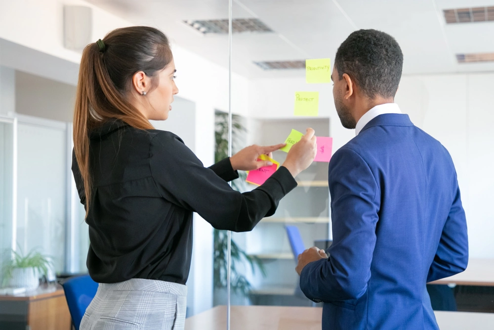 professional-young-colleagues-working-with-stickers-focused-female-worker-sticking-note-glass-focused-businessman-blue-suit-standing-near-her-teamwork-business-collaboration-concept_1000x667